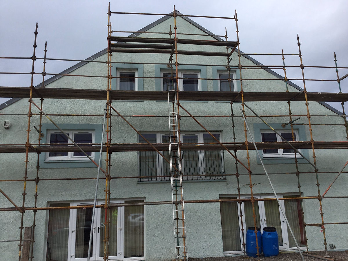 A green-painted house surrounded by scaffolding and water drums during roughcasting work